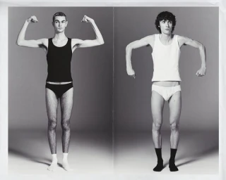 Two male models posing in underwear flex their arms against a grey studio backdrop in a black and white editorial image.
