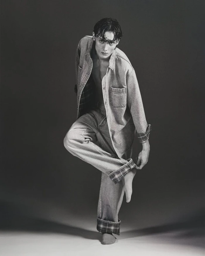 Young male model posing in denim shirt and cuffed trousers against a dark gradient studio background