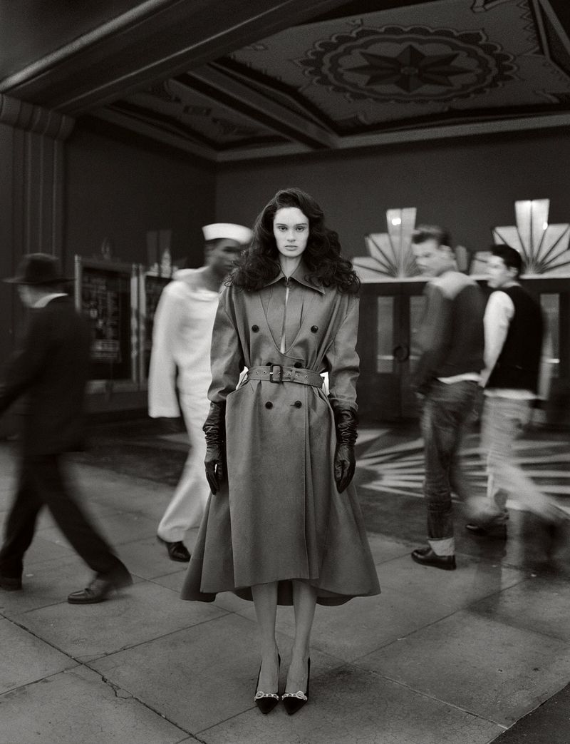 A fashionable woman in a trench coat and heels stands on a patterned floor. Blurred people are in the background of this urban fashion shot.