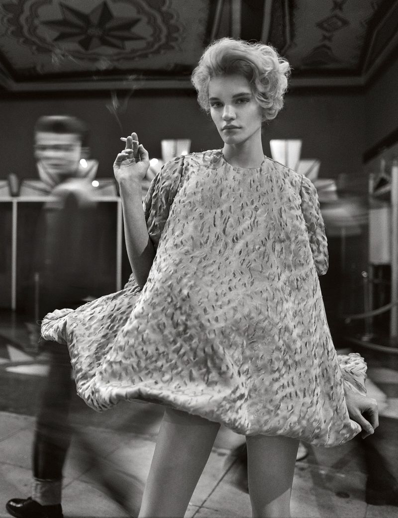 Monochrome shot of a stylish young woman in a textured mini-dress, holding a cigarette, with a blurred figure and patterned wall behind her.