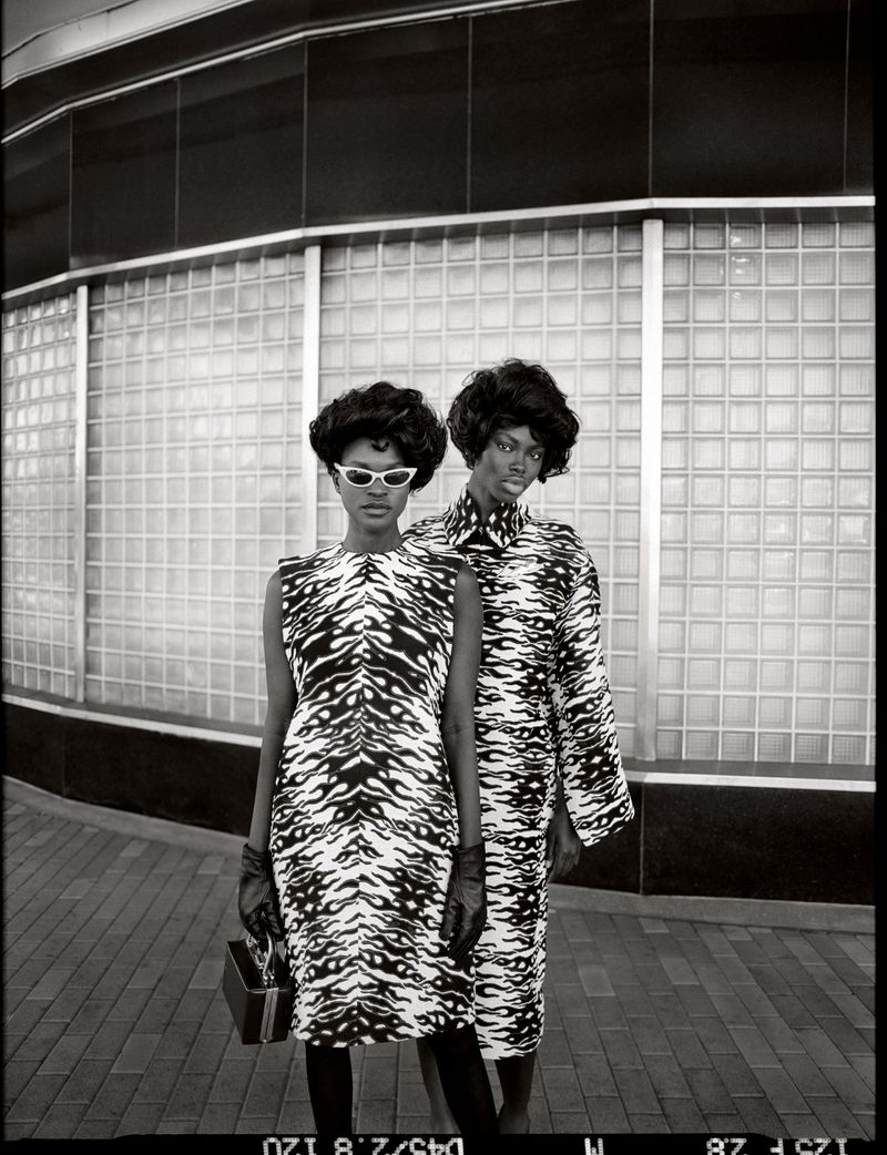Two Black female fashion models pose outdoors in matching black and white tiger-striped dresses and gloves against a tiled wall.