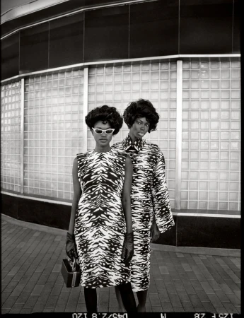 Two Black female fashion models pose outdoors in matching black and white tiger-striped dresses and gloves against a tiled wall.