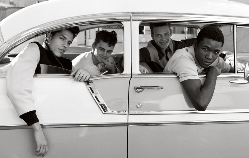 Monochromatic image showcases four young men inside a classic car, leaning out of the windows. Black and white color palette emphasizes detail.