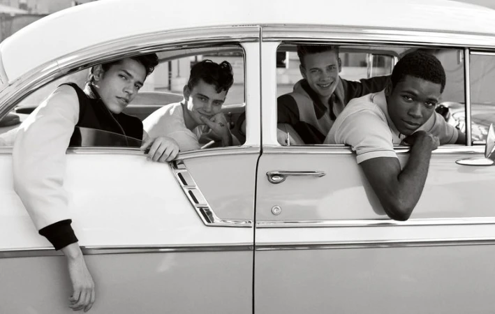 Monochromatic image showcases four young men inside a classic car, leaning out of the windows. Black and white color palette emphasizes detail.