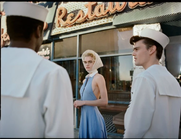 Retro fashion shot of a fair-skinned woman in a blue dress, flanked by two men in sailor uniforms, in front of a diner with neon sign.