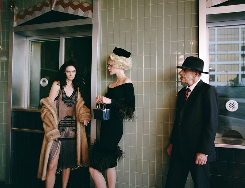 Two women in vintage dresses and a man in a suit stand in front of a building with tiled walls, evoking a classic, retro fashion style.