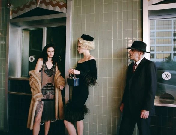 Two women in vintage dresses and a man in a suit stand in front of a building with tiled walls, evoking a classic, retro fashion style.