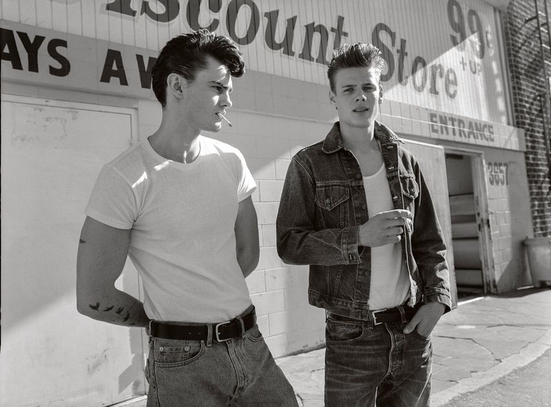 Black and white image of two young men in casual wear standing outside of a store on a city street, one smoking a cigarette.