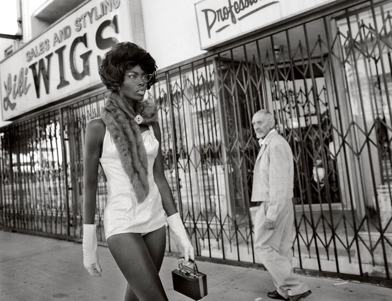 A black and white photo of an African American woman in vintage attire walking past a store with an older man in the background.