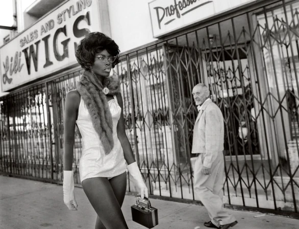 A black and white photo of an African American woman in vintage attire walking past a store with an older man in the background.