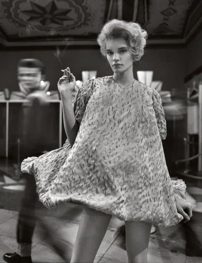 Model in textured mini dress holding a lit cigarette with a blurred figure walking behind in a vintage-styled interior
