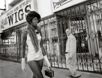Black model walking past a wigs storefront wearing a light-coloured bodysuit, fur stole, and gloves, with a man walking in the background