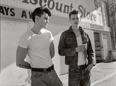 Black and white portrait of two young men standing outside a discount store wearing t-shirts, jeans, and denim jackets.