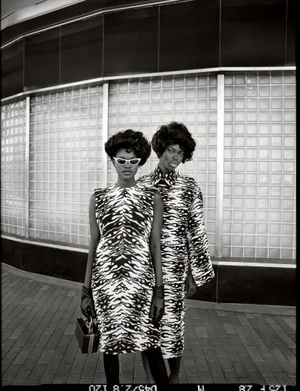 Two models in matching black and white patterned dresses posing in front of a glass block storefront exterior.