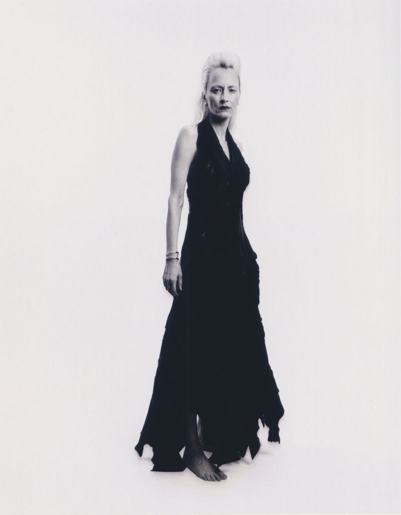 Elegant full-length portrait of a blonde woman wearing a long black gown against a plain white backdrop in a studio environment.