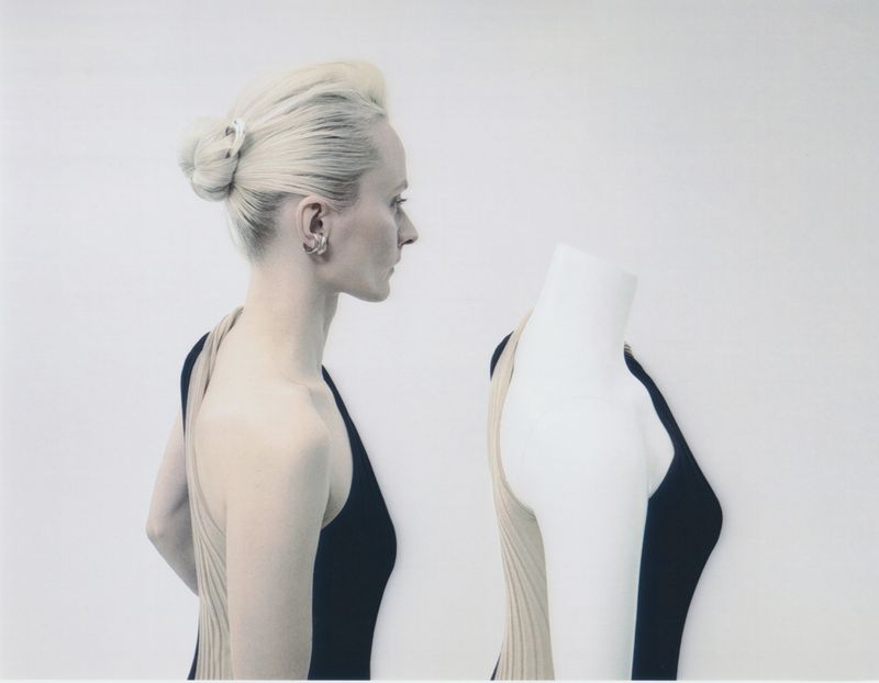Elegant studio shot: profile view of a blonde woman with an updo and a black dress next to a dress form against a white backdrop.