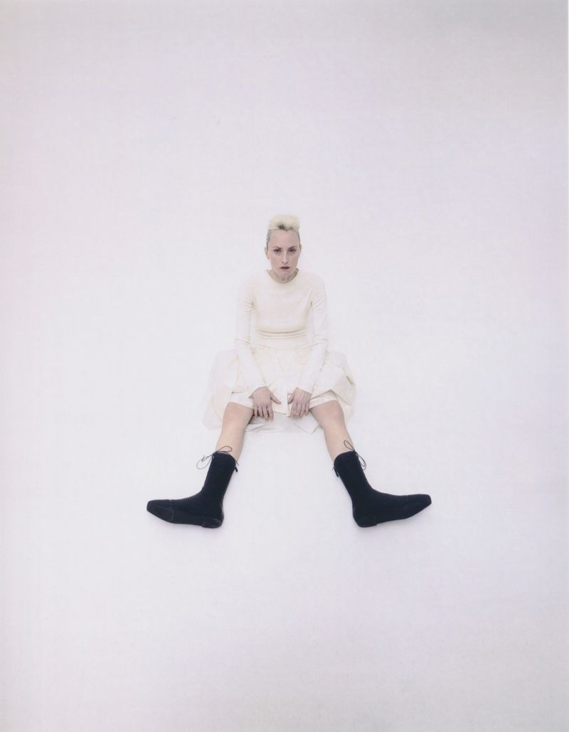 Model with blonde hair seated on the floor wearing a cream dress and black socks against a white backdrop.