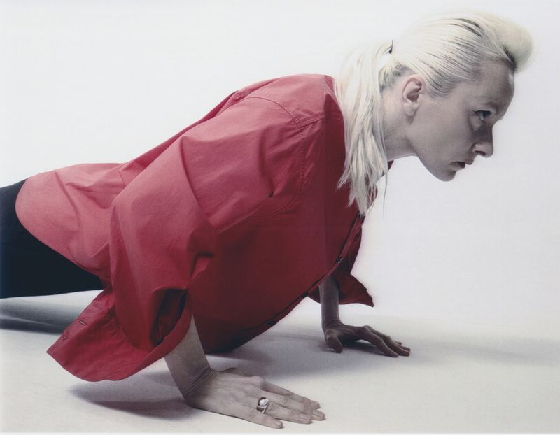 An adult female with pale blond hair is doing a push-up while wearing a red shirt against a solid white backdrop in a studio setting.