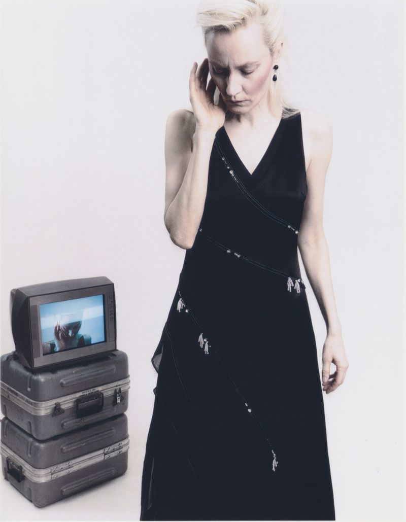 Elegant woman in dark dress near stacked vintage suitcases and an older television set against a minimalist background.
