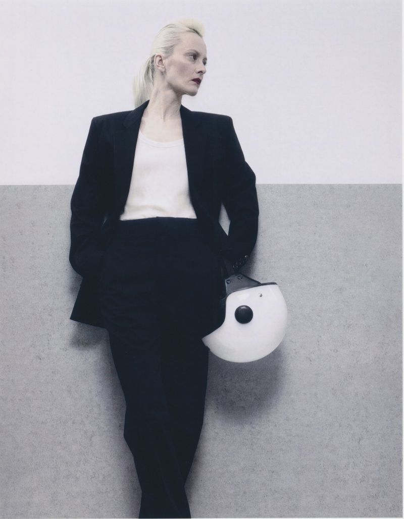 A stylish woman in a blazer poses against a two-tone background, holding a white helmet. Fashion editorial photography, indoor setting.