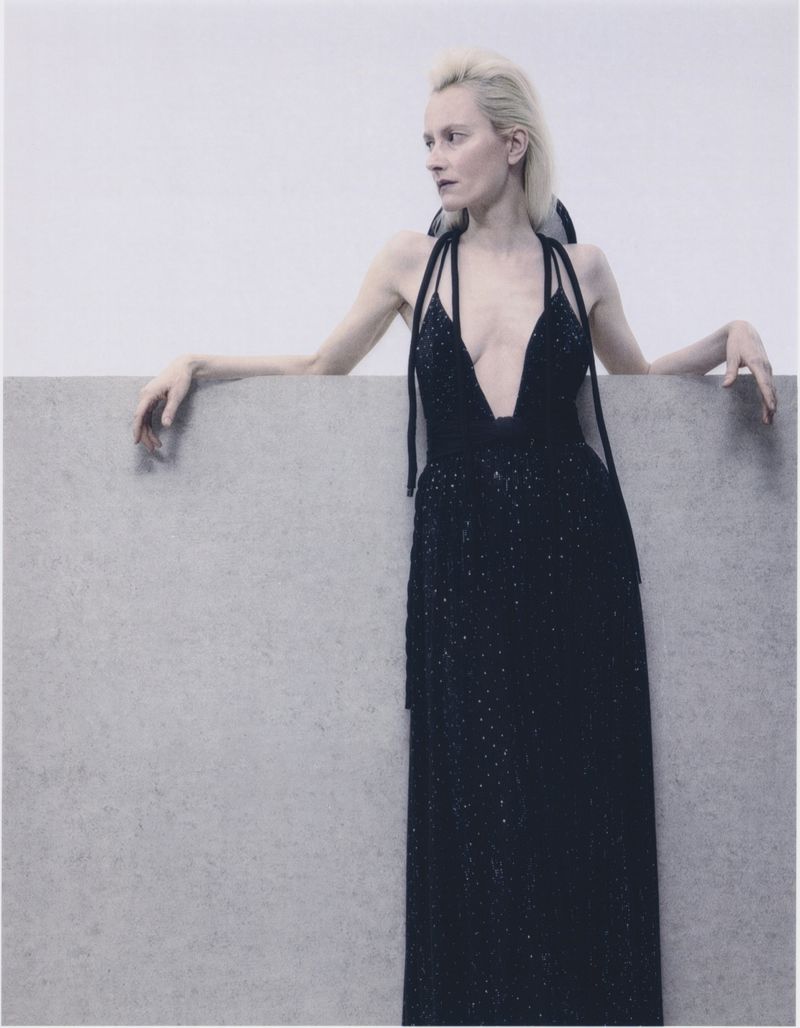 Elegant woman in a black dress with a plunging neckline poses gracefully against a simple gray backdrop, looking off to the side.