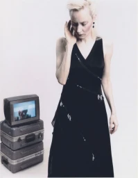 Model in a black V-neck dress standing beside a small vintage television resting on stacked grey cases.