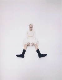 Model with blonde hair seated on the floor wearing a cream dress and black socks against a white backdrop.