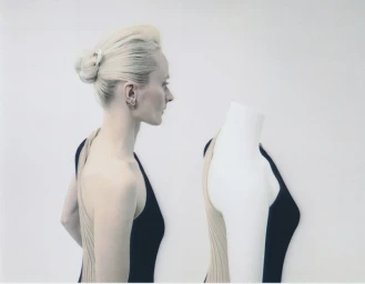 Profile of a blonde woman standing next to a headless white mannequin against a plain white background