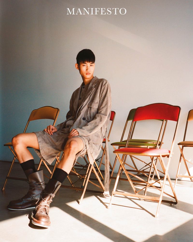 Asian male fashion model poses seated on a folding chair wearing a grey button-down shirt and shorts with high boots in a studio.