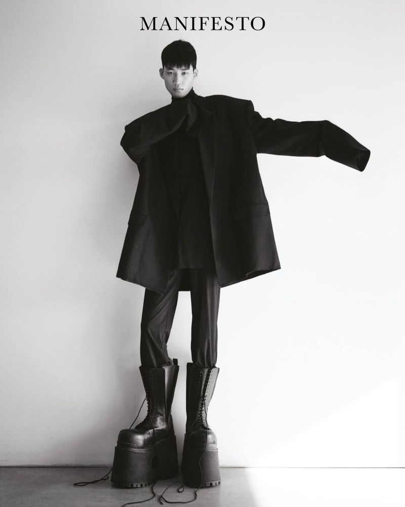 Person in black turtleneck, trousers, coat, and platform boots poses against a white wall, wearing fashion from Manifesto Magazine.