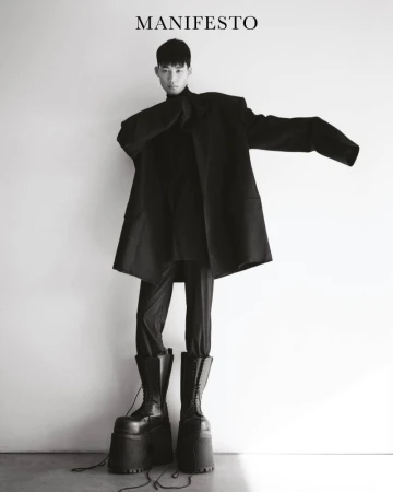 Person in black turtleneck, trousers, coat, and platform boots poses against a white wall, wearing fashion from Manifesto Magazine.