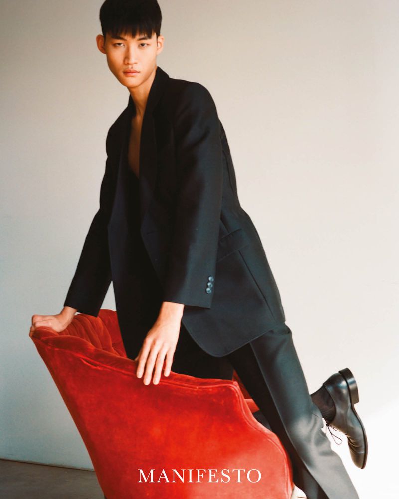 Stylish young man in an oversized blazer and dress shoes leaning against a red velvet chair in a fashion editorial shot.