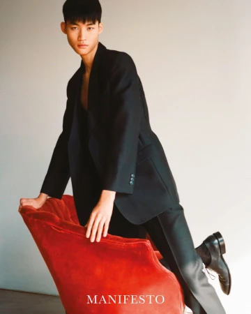 Stylish young man in an oversized blazer and dress shoes leaning against a red velvet chair in a fashion editorial shot.