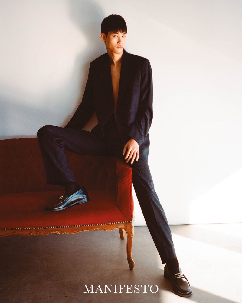 Asian male fashion model in a black suit poses on a red vintage sofa against a white wall, showcasing contemporary style.
