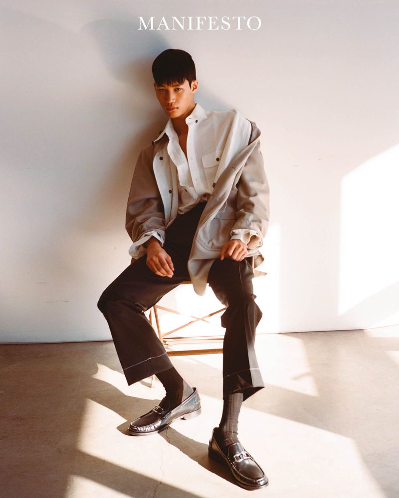 Asian male model in casual wear sitting on a chair in a photo studio with natural light and visible shadows, fashion editorial shot.