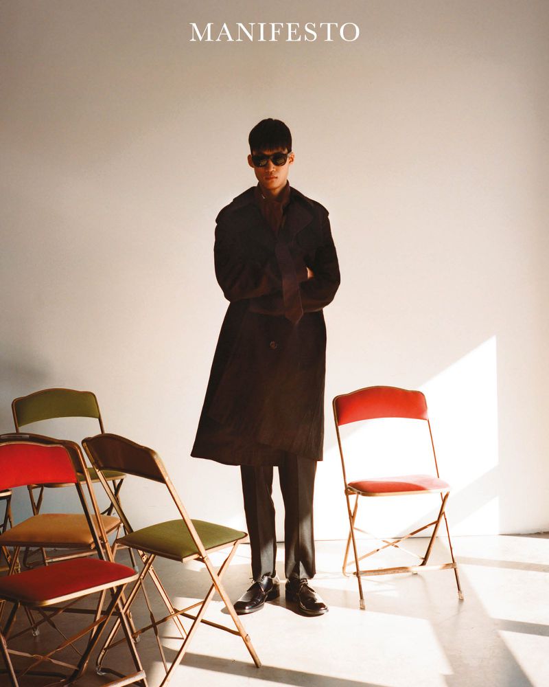Fashionable man in sunglasses and trench coat posing among colorful folding chairs in a brightly lit, neutral indoor setting.