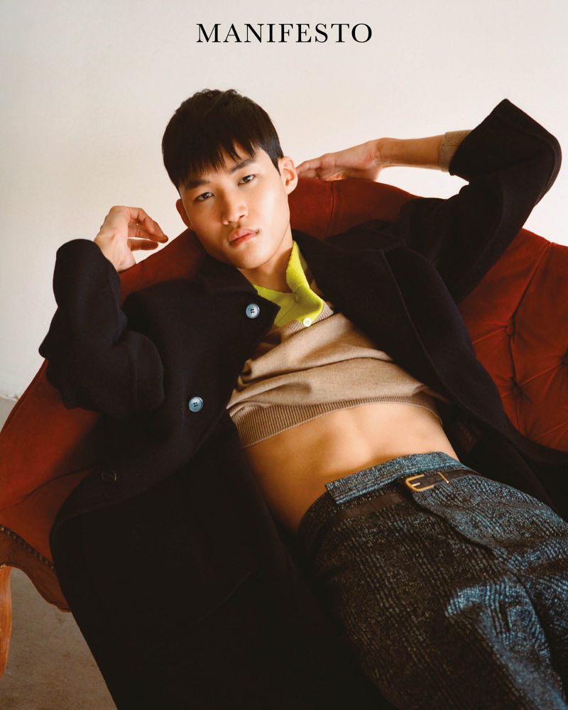 Portrait of a young Asian male fashion model wearing a black coat, beige top, and patterned pants, posing on a red armchair.