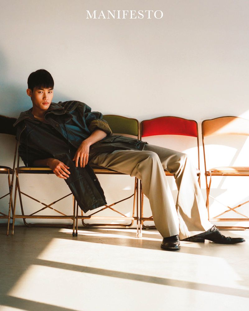 Asian male model reclining across a row of folding chairs wearing a dark jacket and trousers against a white wall.