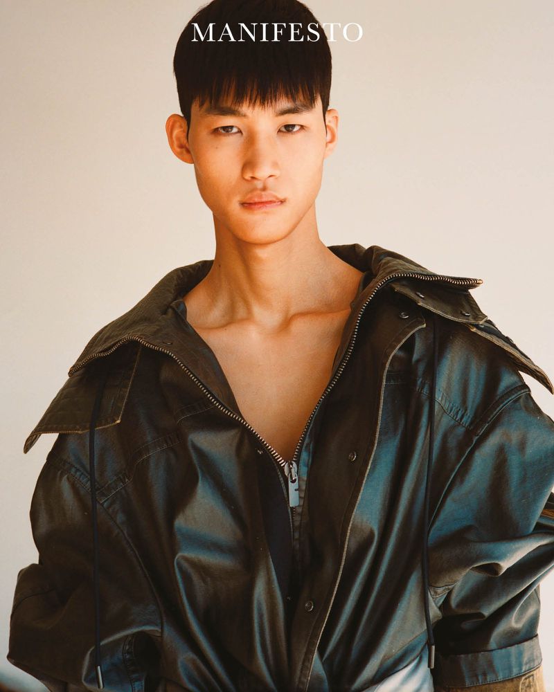 Portrait of a young Asian man in a black jacket with a silver zipper, posing indoors against a neutral wall with the word 'MANIFESTO' above him.