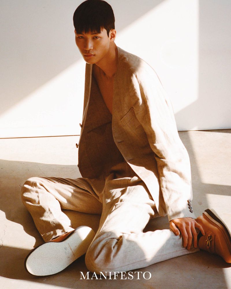 An Asian male model poses indoors wearing a tan blazer and pants, sitting on the floor, with a light spot highlighting his face.