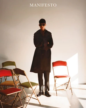 Model wearing dark sunglasses and a long coat standing amid several folding chairs in a light studio setting.