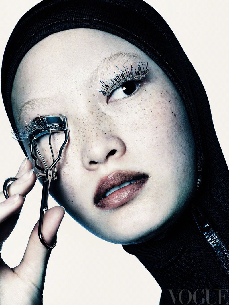 Close-up studio shot of a woman with light skin and freckles using an eyelash curler; she has bleached eyebrows and long false eyelashes.