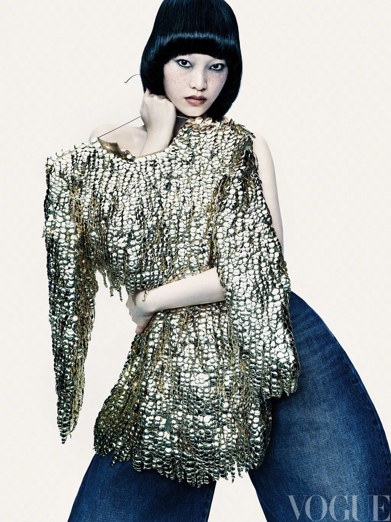 A woman with an asymmetrical bob hairstyle poses in an off-the-shoulder gold metallic top and blue denim wide leg pants in a studio shot.
