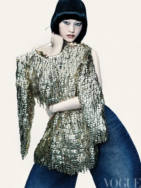 A woman with an asymmetrical bob hairstyle poses in an off-the-shoulder gold metallic top and blue denim wide leg pants in a studio shot.