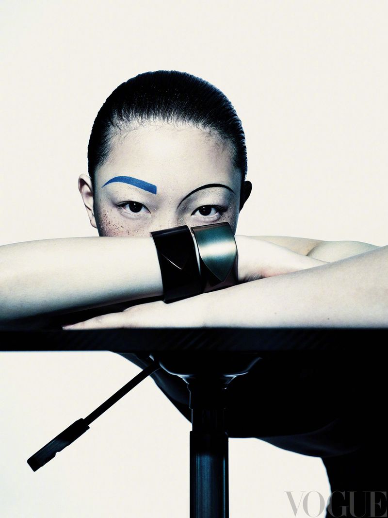 Close-up fashion shot of an Asian woman with unique blue eyebrow and metal cuff bracelet, gazing intensely at the camera with neutral expression.