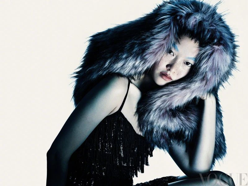 A woman with light skin and freckles poses with a large, furry hat in a studio setting for a fashion shot with the 'Vogue' logo visible.