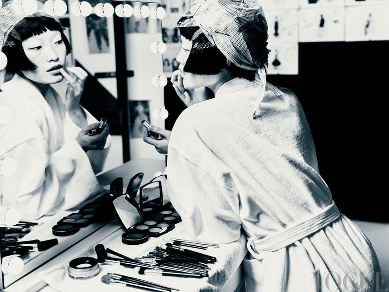 A woman in a robe and head covering applies lipstick in front of a mirror in a black and white fashion photography style.