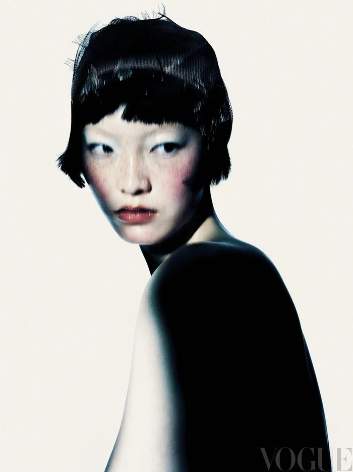 Close-up fashion portrait of an East Asian model with short dark hair wearing a mesh cap against a bright white background.