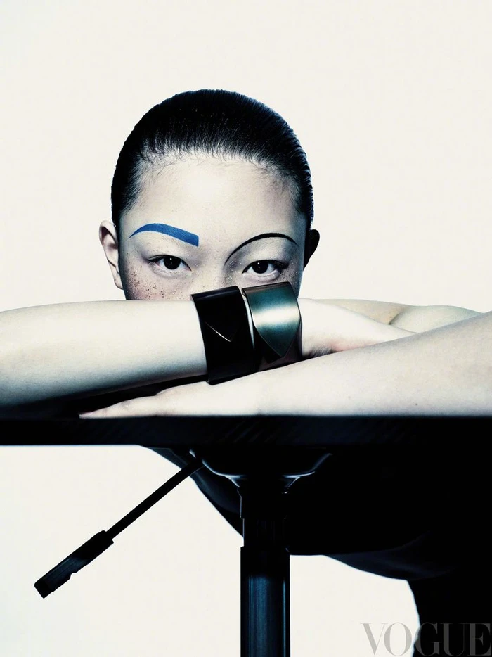 Close-up portrait of model with bold blue eyebrow makeup resting arms on table against white background