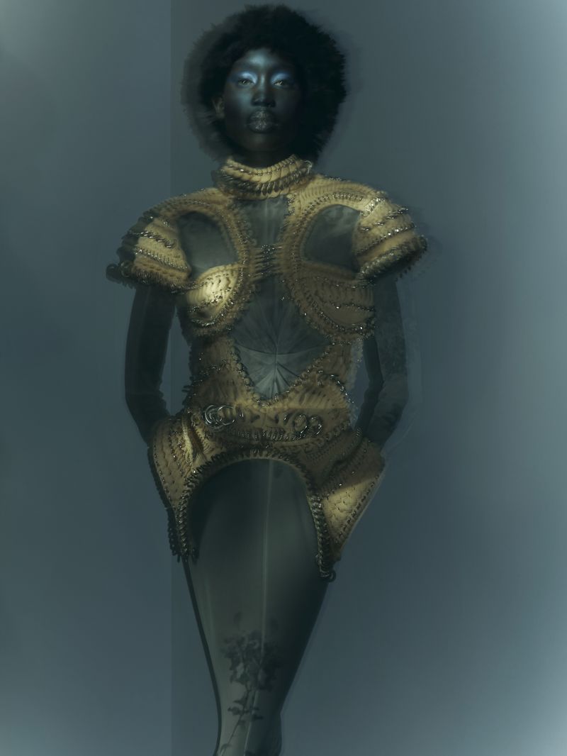 An African American woman with an afro hairstyle poses in a golden, beaded, sculptural couture outfit against a soft, blue-toned studio backdrop.